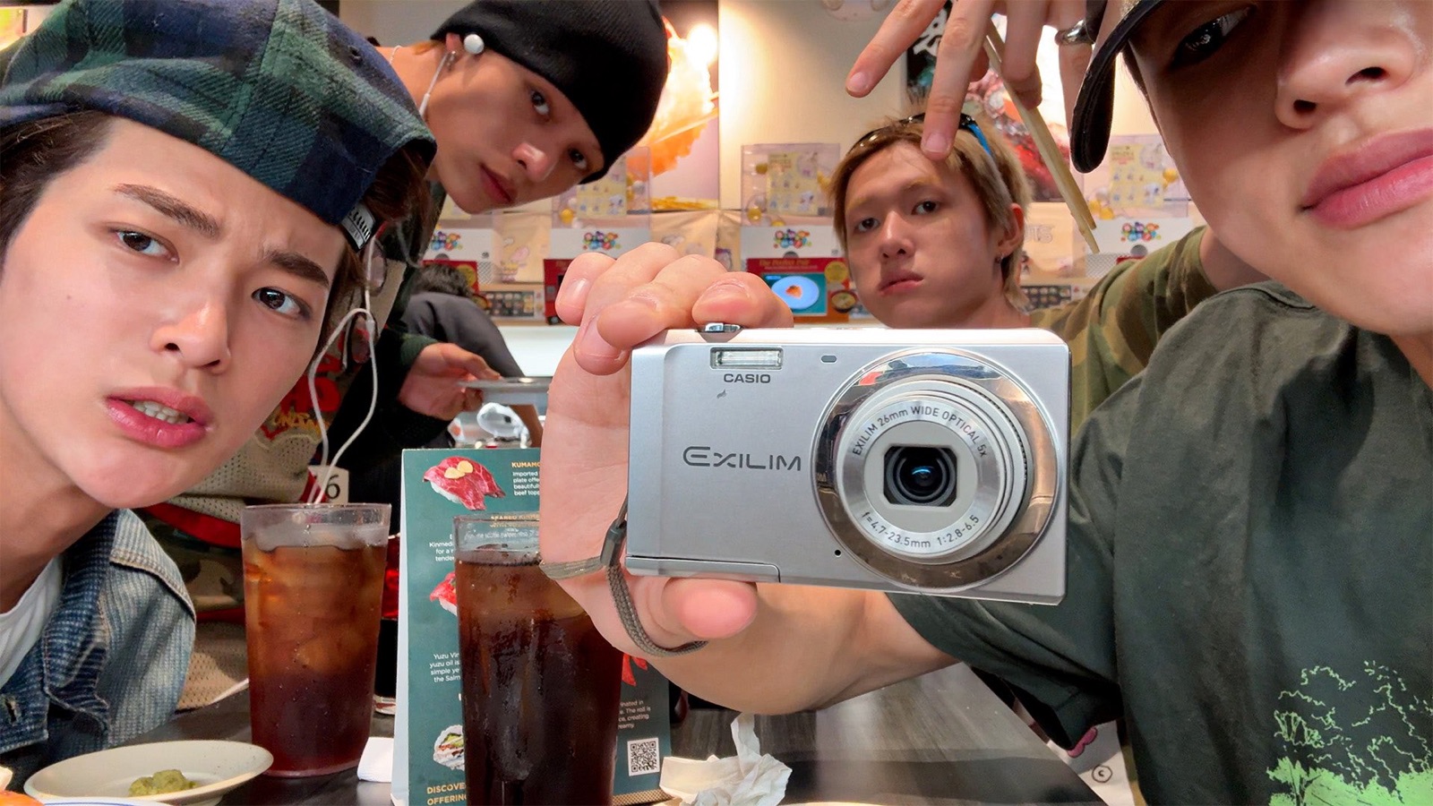 Four members of CORTIS crowded around a silver Casio camera at a sushi restaurant